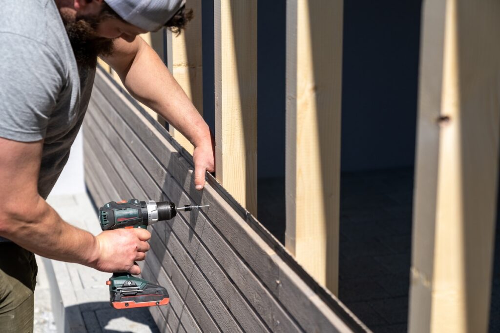 A tough construction worker applies wooden siding to a garage and drills screws