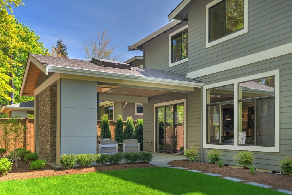 Large modern grey new house with back porch from back yard with green grass and nice landscaping.