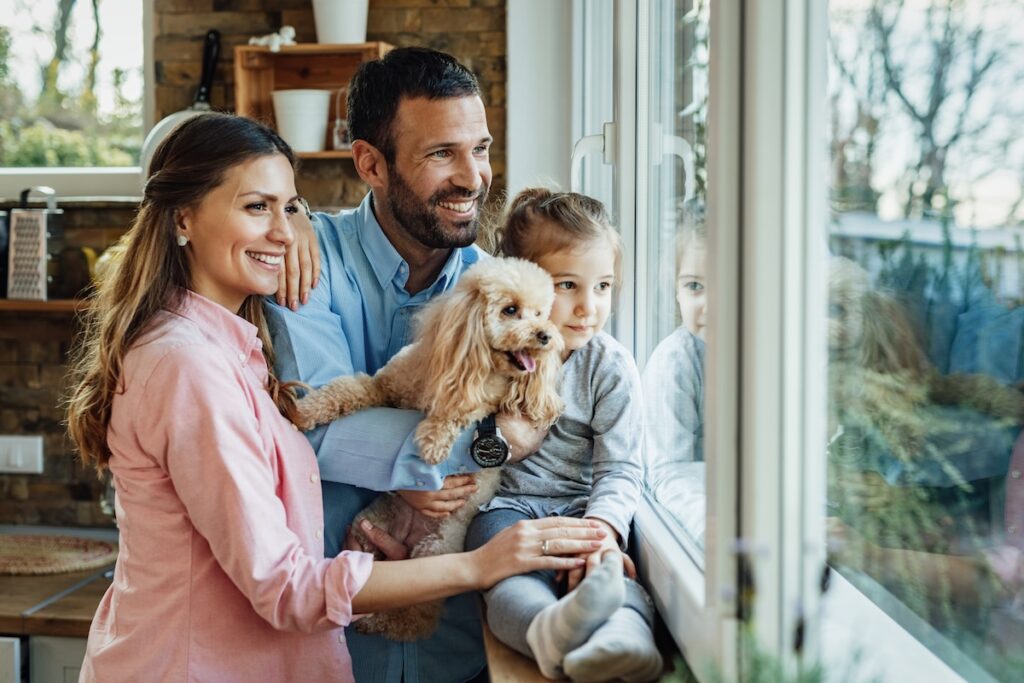 Young happy family and their dog relaxing by the window at home and looking through it