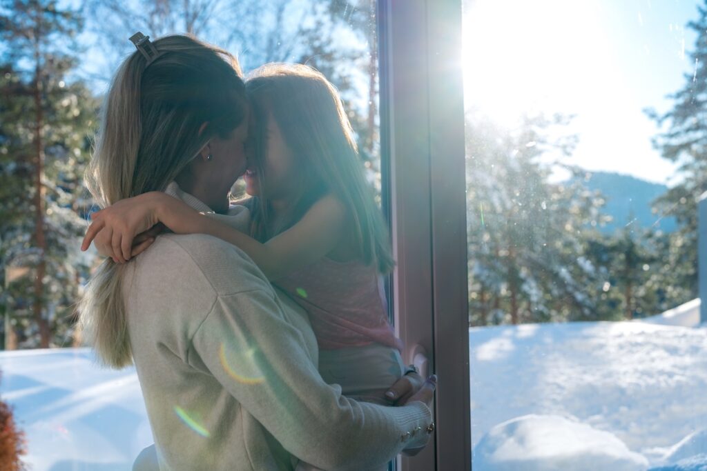 Mother embracing her daughter while looking out at a snowy landscape through a large window