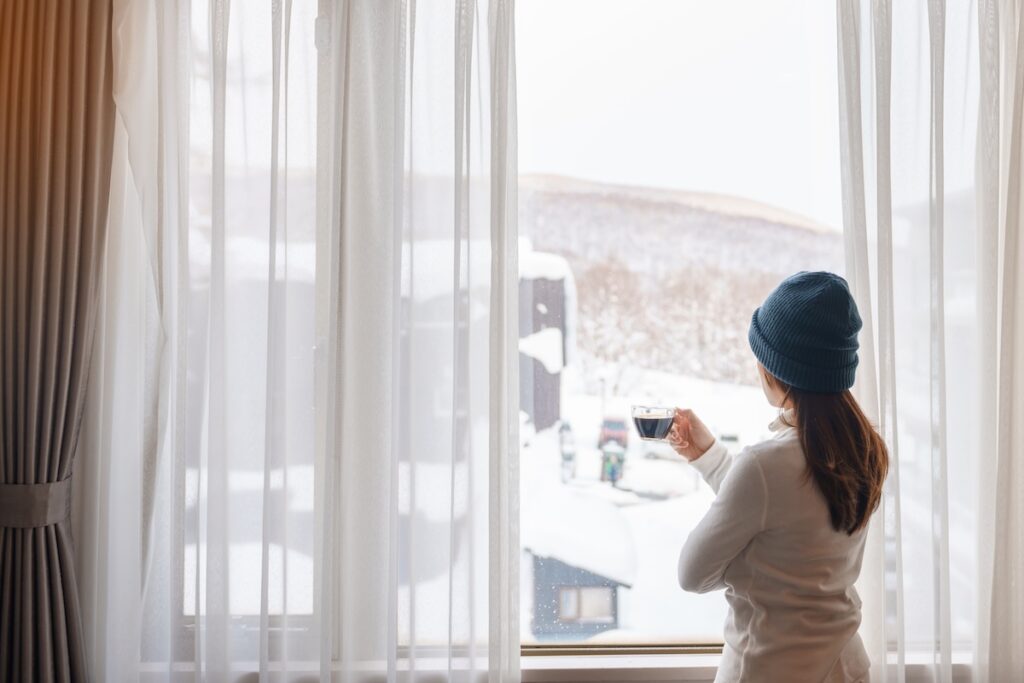 young woman in sweater with cup of coffee looking through the window 